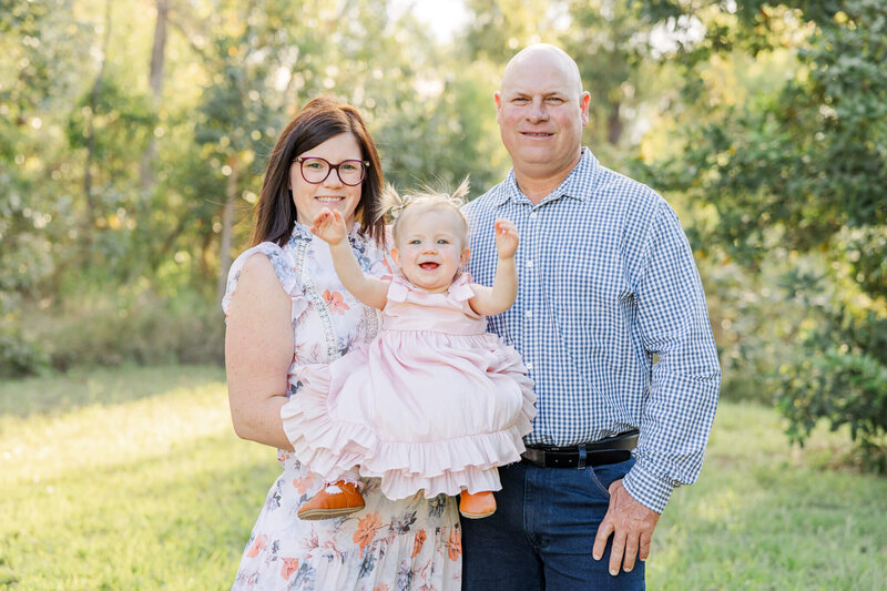 Gorgeously dressed parents, standing in an orchard, smiling at the camera, holding a toddler baby girl dressed in a pink dress. Little girl waves happily at the camera. 