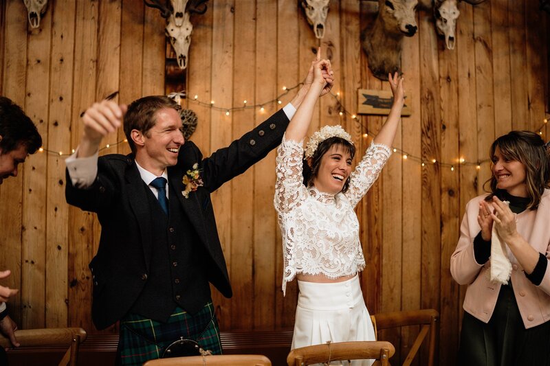 A bride and groom are photographed in Mar Lodge’s ballroom in Aberdeenshire during their wedding reception. The wide photo pictures them holding their hands in the air with joy as guests are photographed all around them cheering. The atmosphere is warm and inviting. 