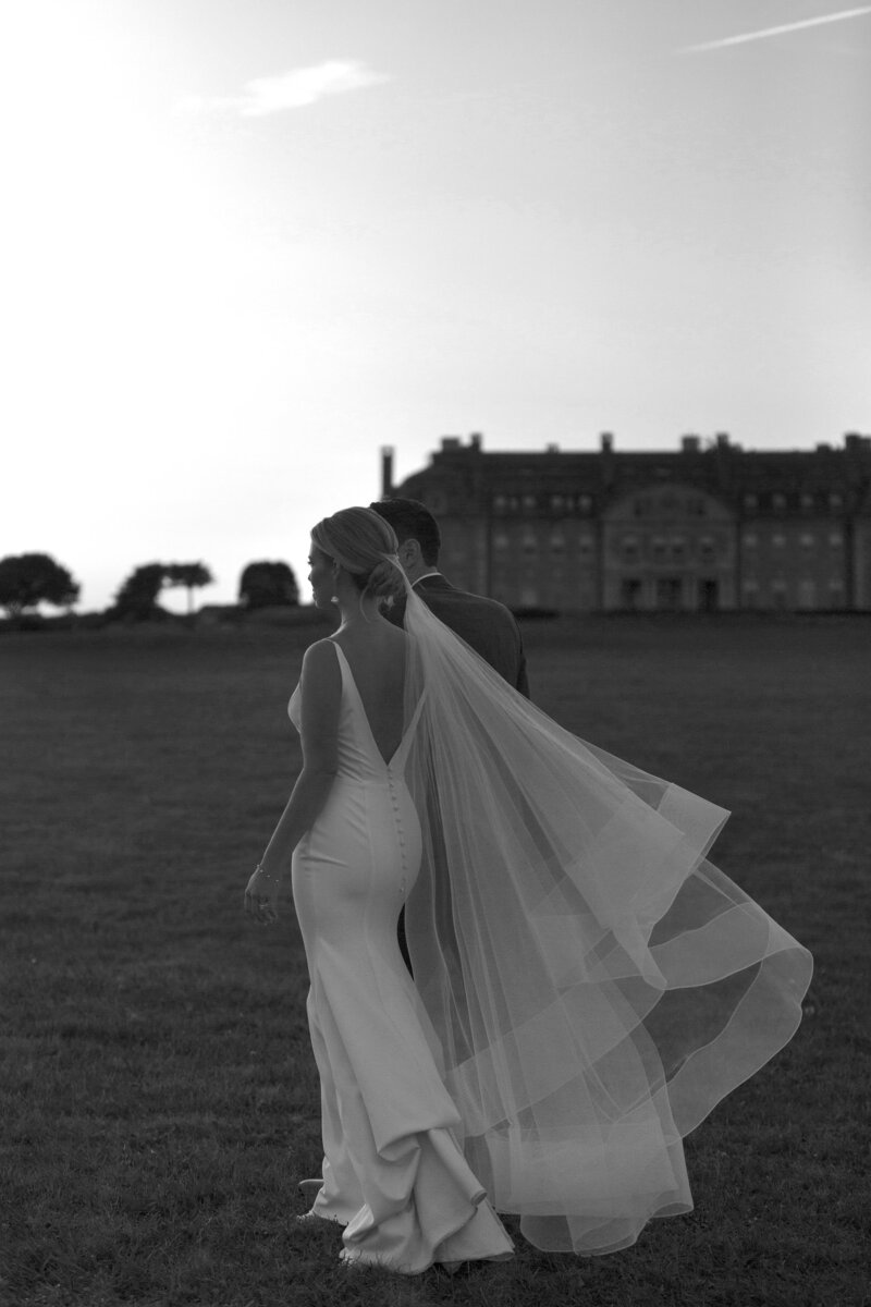 Black and white photo of bride facing away from camera with veil blowing in the wind. Deborah Zoe.