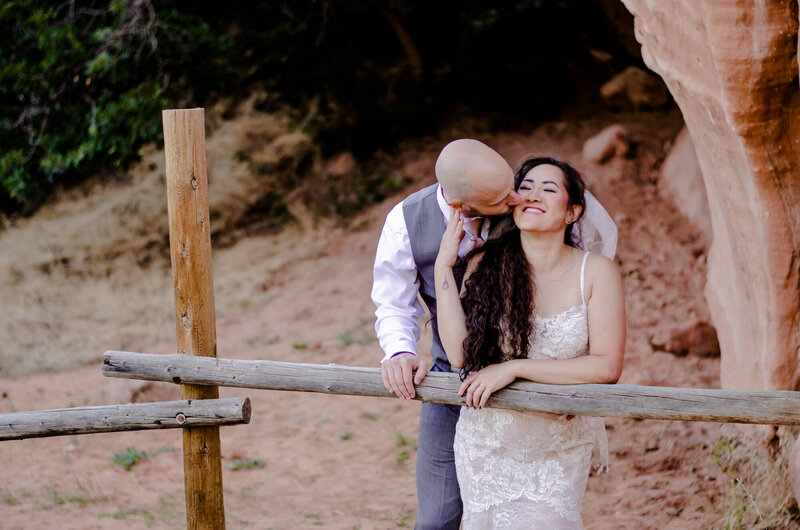 bride being kissed from behind by her groom. Standing against a wooden fence near red rocks