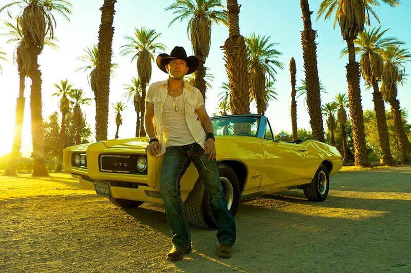 Country Music Photo Aaron Pritchett standing in front of yellow convertible GTO surrounded by palm trees