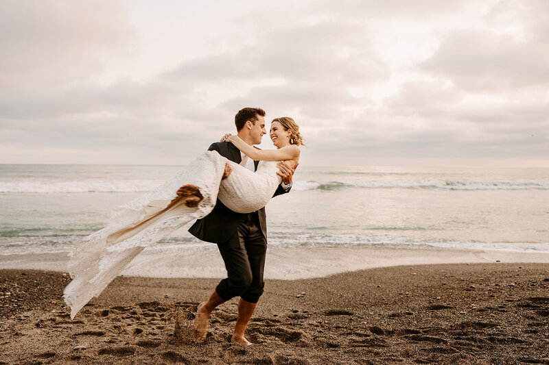 groom holds bride on beach