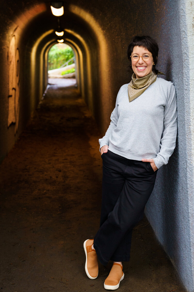 A woman associate professor with tenure leans casually against the wall of a tunnel with her hands in her pocket and one leg crossed over the other. She is smiling and has short, dark brown hair. She is wearing a grey sweatshirt, black pants, and a green neck scarf. There are lights on the ceiling of the tunnel and you can see greenery on the other side.