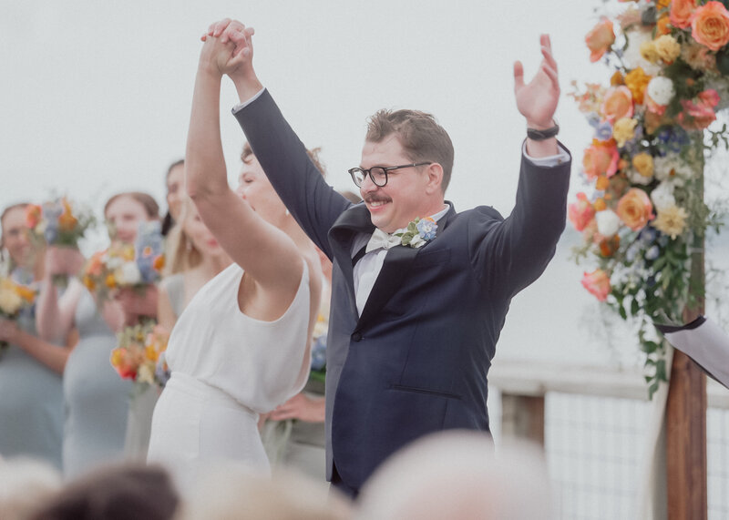 A couple in an outdoor wedding holding  their hands in the air with a audience clapping  around them.  There is a bed of flowers behind them