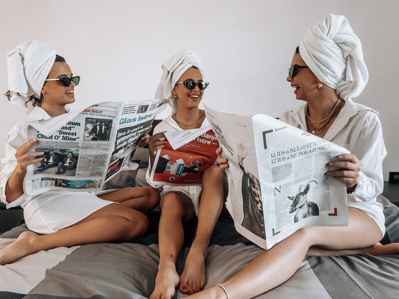 3 women relaxing in robes and towels, reading newspapers