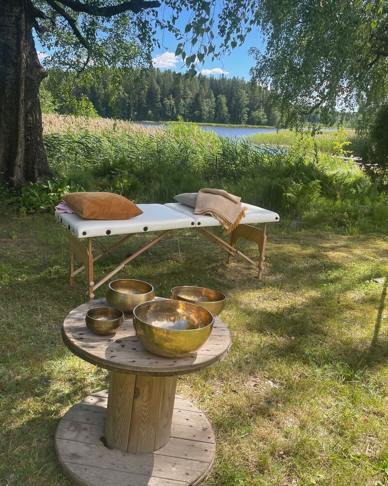 Hoitopöytä valmiina äänimaljahoitoihin ja kehohoitoihin. Kuvassa näkyvä hoitopöytä tarjoaa mukautetun ja rauhoittavan ympäristön rentoutumiseen ja sointukylpyihin. Taustalla upea suomen luonto
