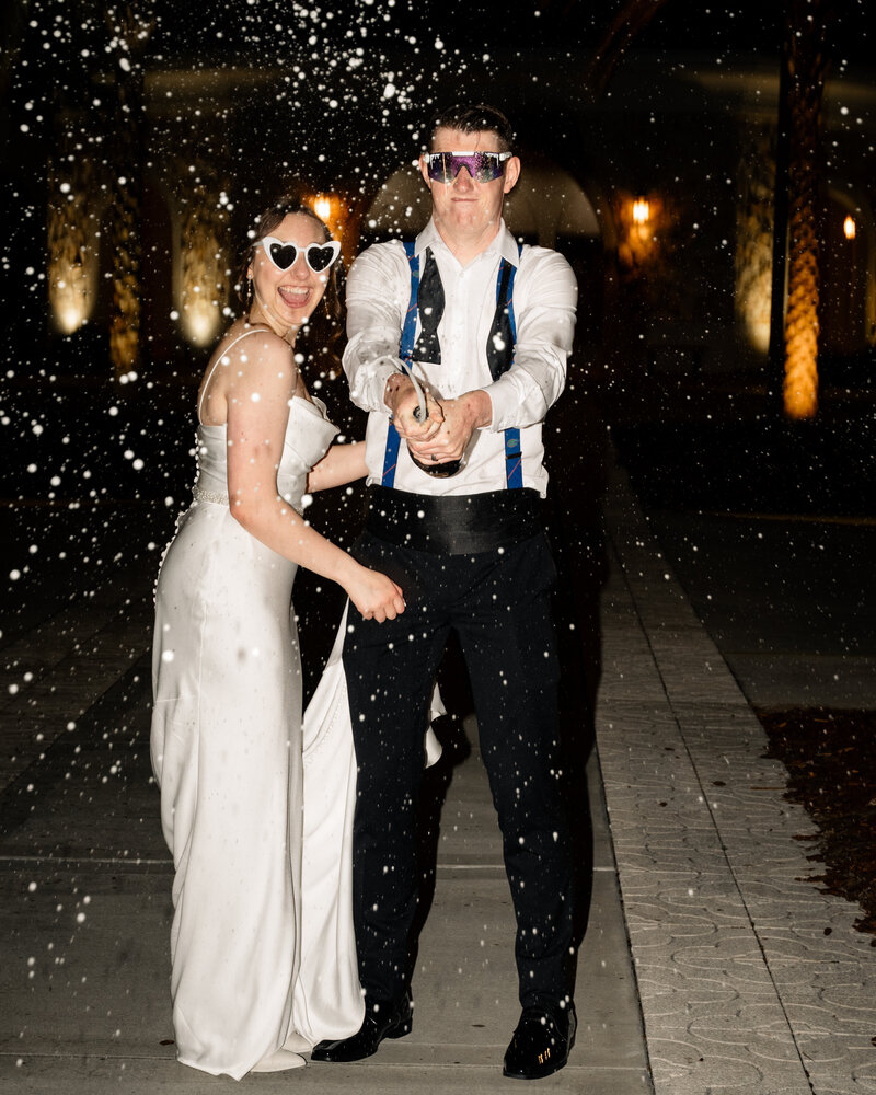 A bride and groom do a late-night champagne pop at their Catholic wedding reception at St. Anastasia Catholic Church in St. Augustine, Florida. They are wearing sunglasses and laughing