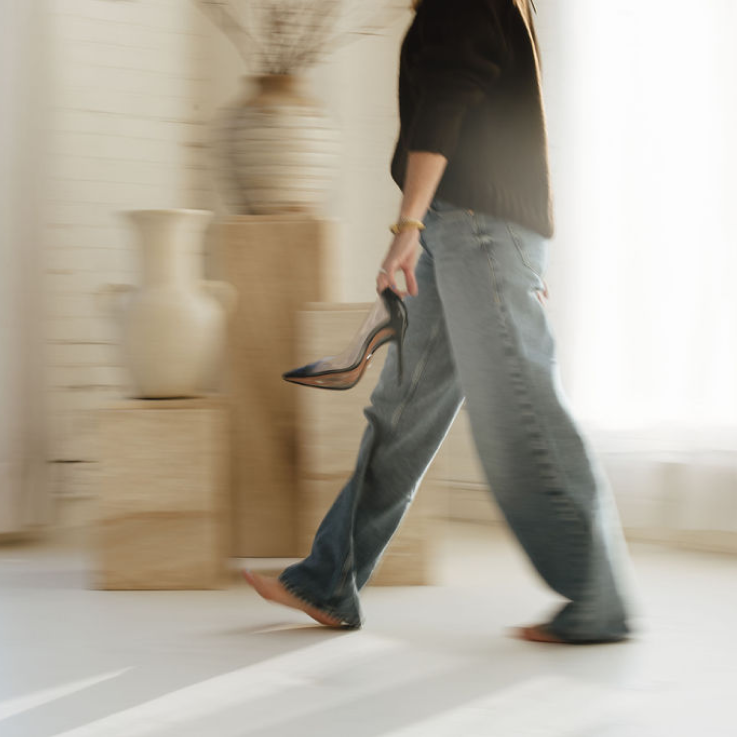 A woman walking barefoot across a living room space with high heels in her hand.