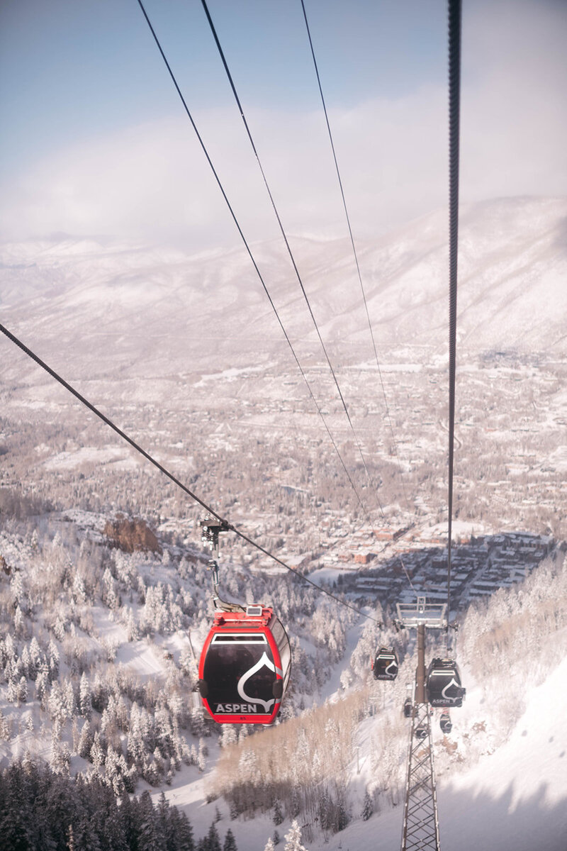 Image shot from a gondola ascending aspen mountain looks down at the snowy town of aspen. Other gondola cars are in the foreground.
