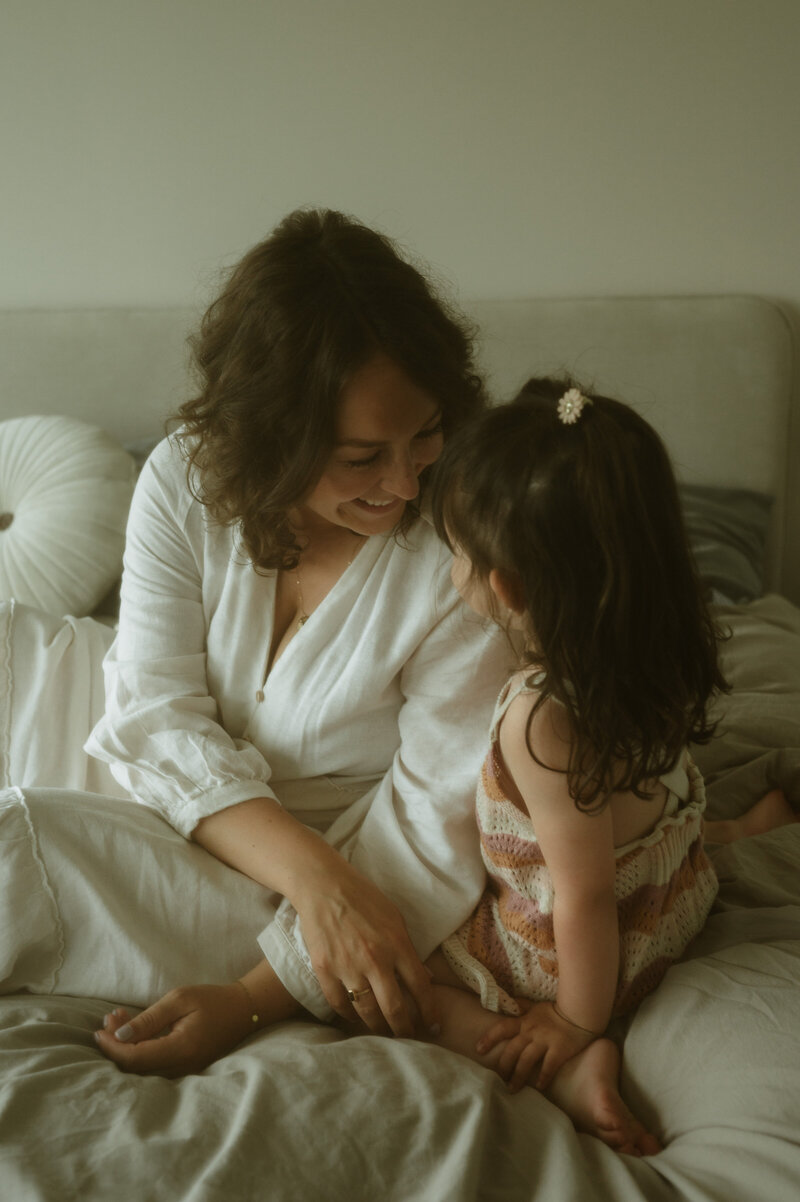 Mama en haar dochter op het bed met kleding in natuurlijke tinten die elkaar liefdevol aankijken.
