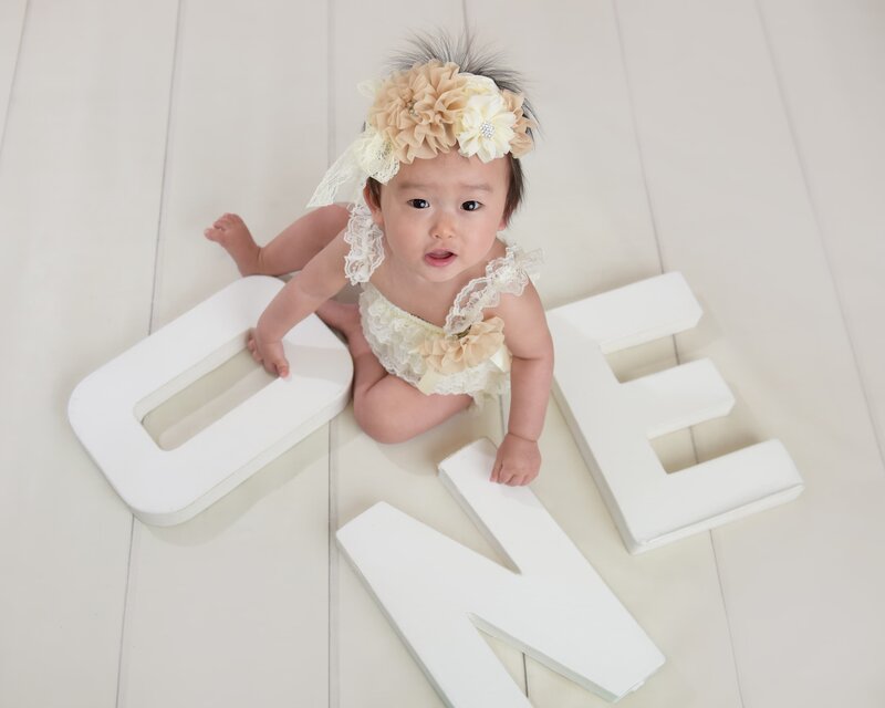 A baby girl wearing a delicate cream romper with floral accents and a matching headband, posing on the floor next to white "ONE" letters.