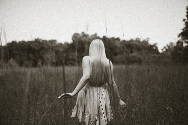 Emotive black and white senior portrait of a girl walking away from the camera with her hands brushing the tips of the grass. 