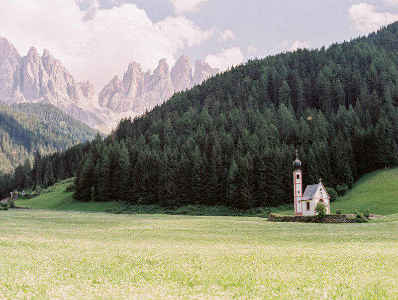 Small church building in front of forest and dolomites mountains with open prairie