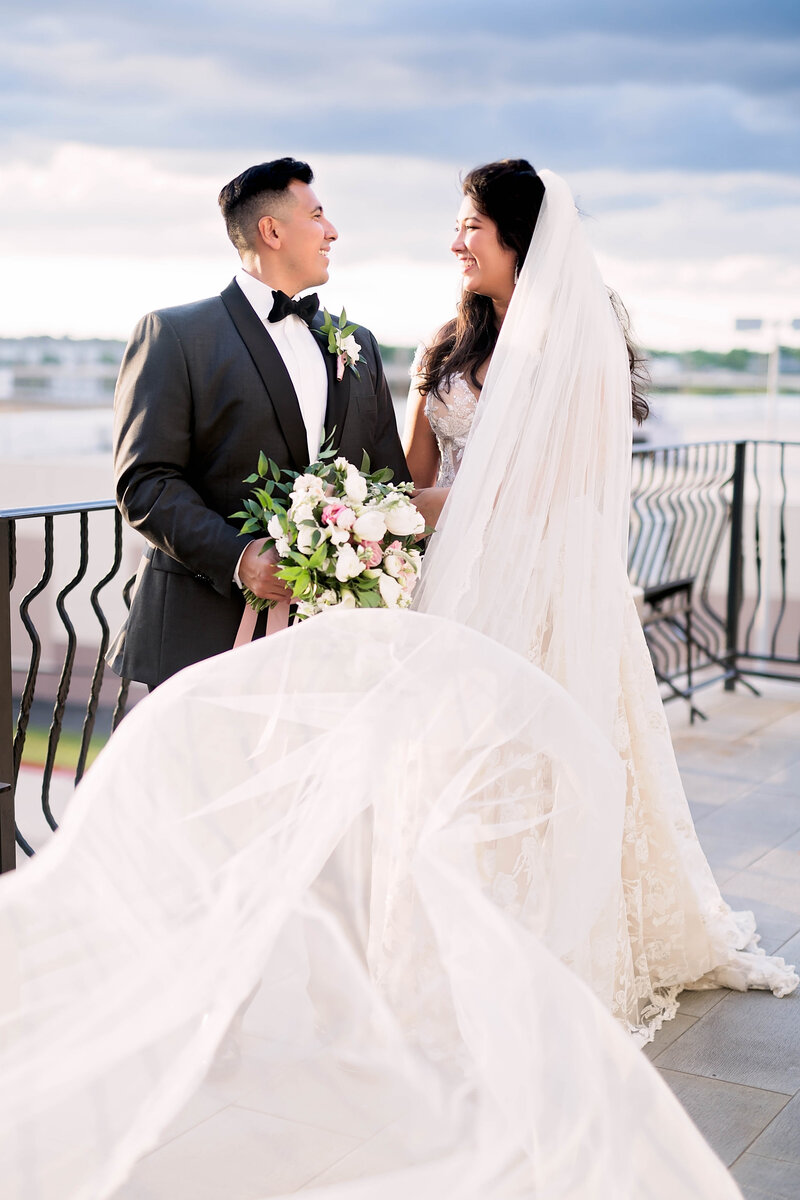Bride and groom smile at each other on a rooftop overlooking a river. The bride's veil flows towards the camera.