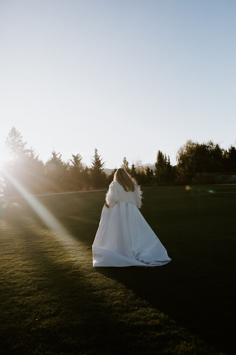 Image of a bride walking away from the camera in a beautiful, full white skirt with a "fluffy" jacket on for warmth. The sun is setting and creating a golden glow to the image.