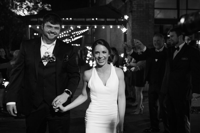 Black and white photo of bride and groom exiting the reception. Family and friends surrounding as bride and groom leave wedding reception. Couple smiling and holding hands.
