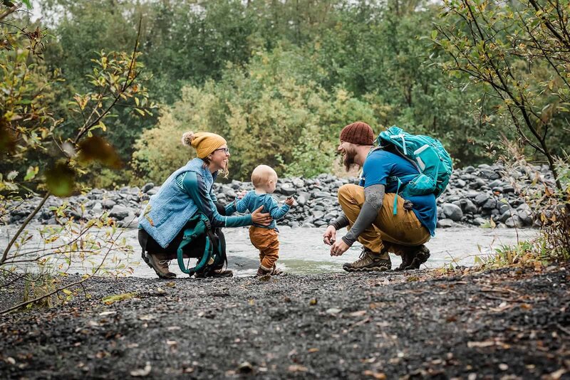 A couple crouches with their young child standing between them next to a rushing river during their private family excursion on day two of their 3 day luxury elopement in Alaska.