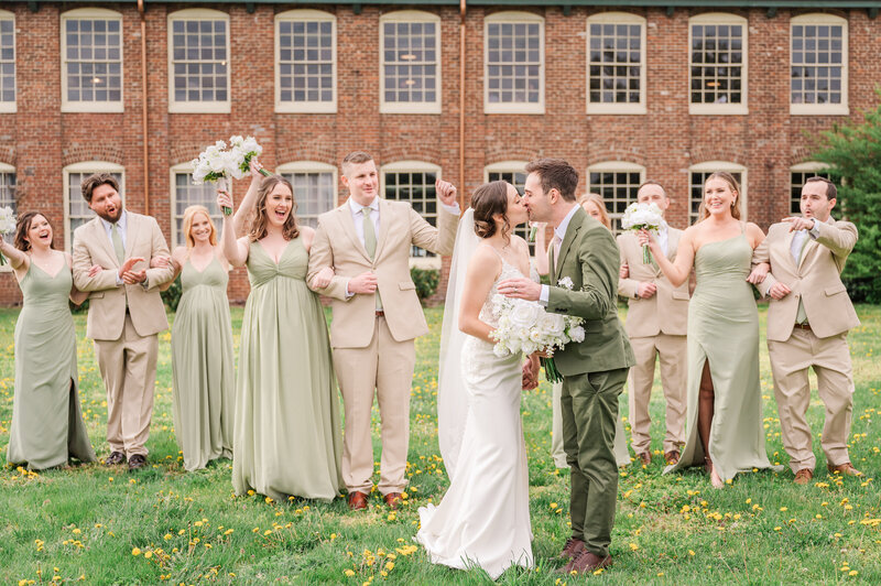 A bride and groom kissing in front of The Woolen Mill during their spring wedding surrounded by their cheering wedding party in sage green and tan by JoLynn Photography