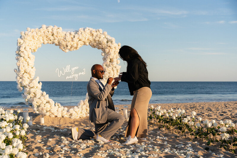 Beach proposal with a man on one knee in front of a white floral heart arch and ‘Will you marry me?’ sign, styled by Yours Truly, a wedding planner and designer in New Jersey.