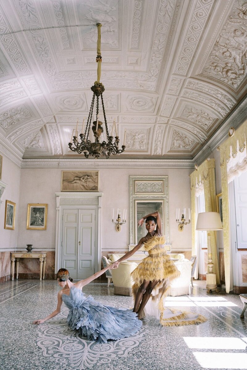 A bride in a voluminous white wedding dress poses gracefully against a stone railing in an elegant, arch-filled corridor in the Louvre, Paris. The dramatic gown trails behind her on the checkered floor, its beauty perfectly captured by luxury international wedding photographer Andreas K. Georgiou.