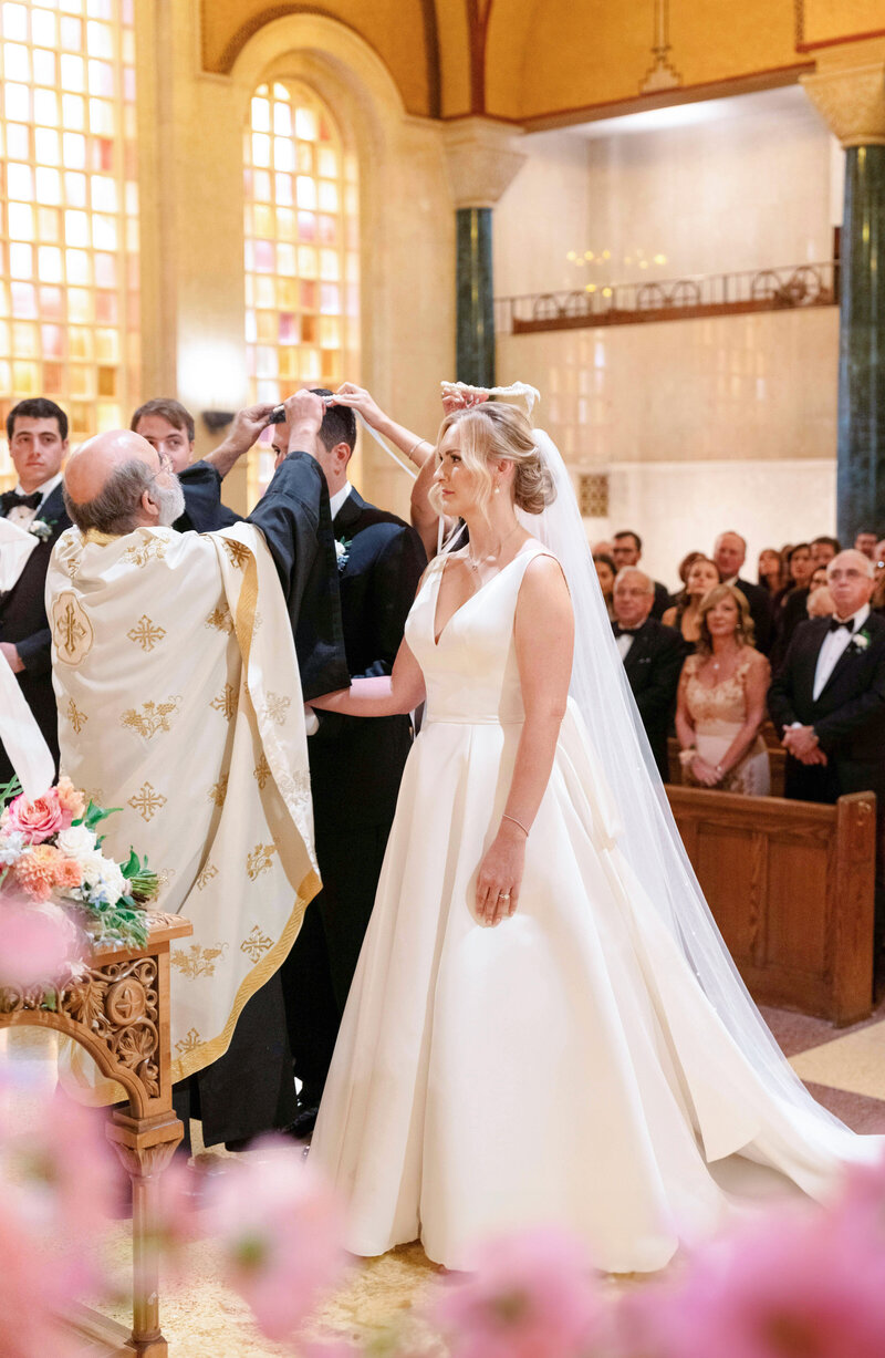 A bride and groom stand in a church during their wedding ceremony. The bride wears a white dress and a veil. A priest in ornate robes performs the ceremony while guests seated in pews look on. The church is decorated with flowers.