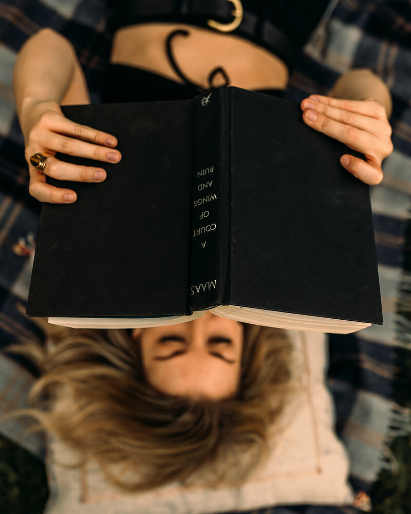 Lauren Hughes lays on her back on a plaid blanket. She's holding the book, a Court of Wings and Ruin up and she's wearing a gold tiger's eye ring.