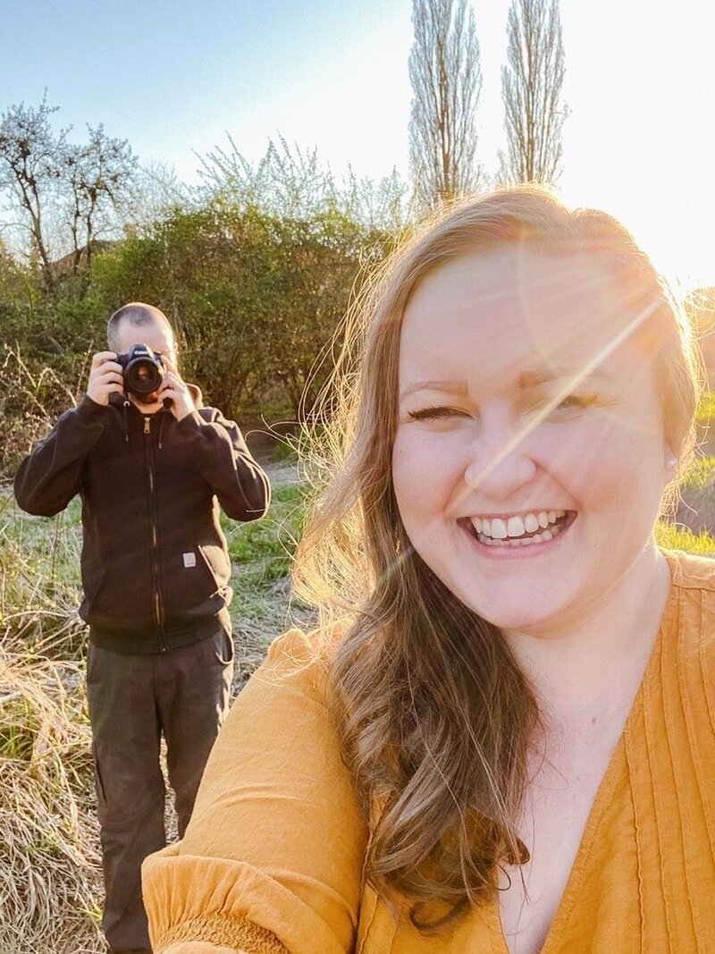 Laura-Anne smiling in a selfie while her husband holds up a camera in the background