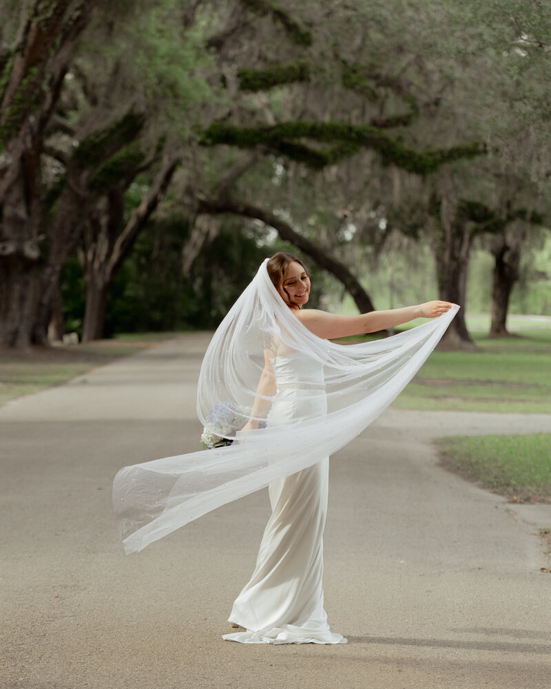 A bride twirls around with her veil under live oaks with spanish moss at St. Ambrose catholic church in St. Augustine, Florida. She is full of joy after her catholic wedding Mass.
