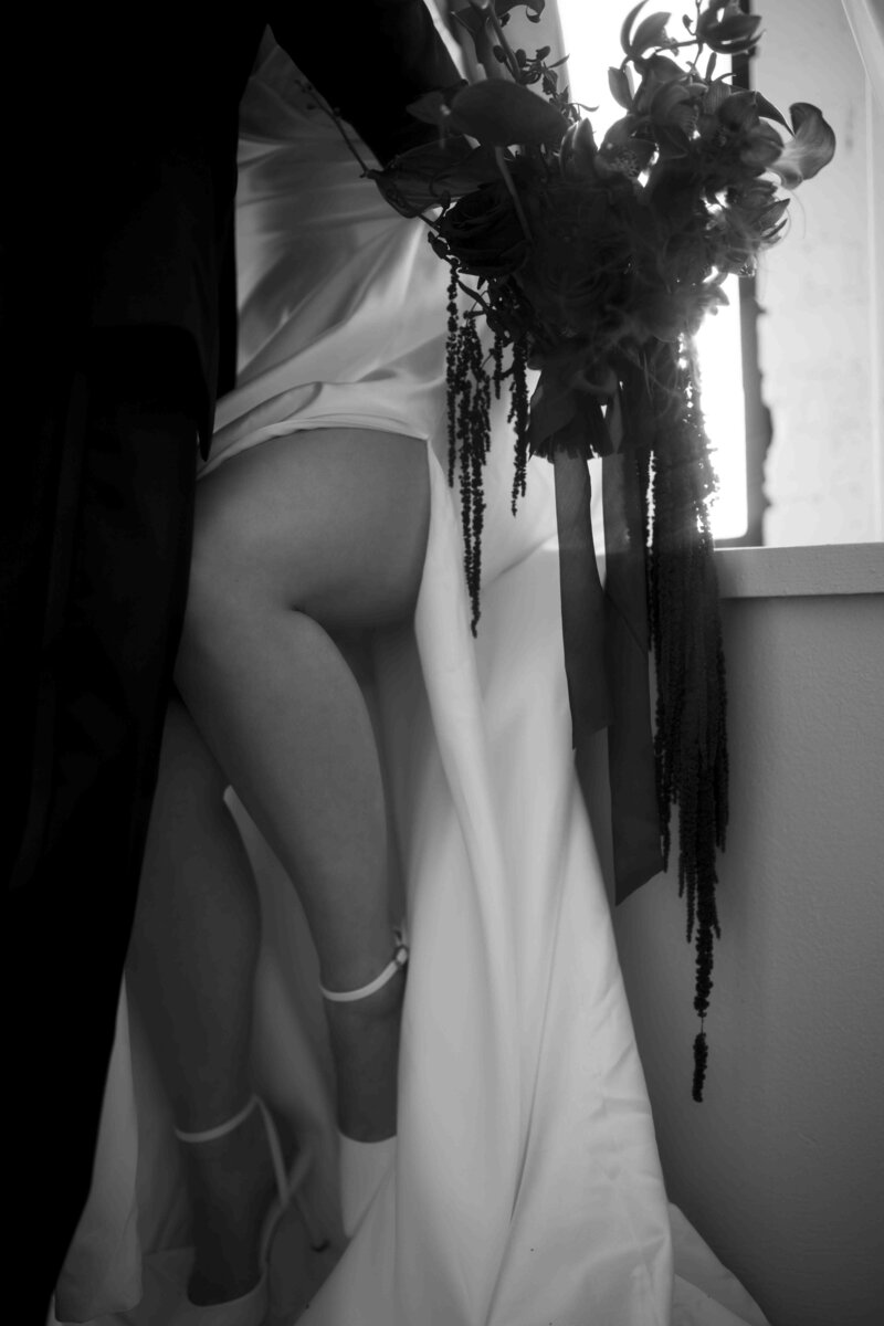 Bride and groom leaning against a window sill, close up of legs, dress, and the flowers she is holding