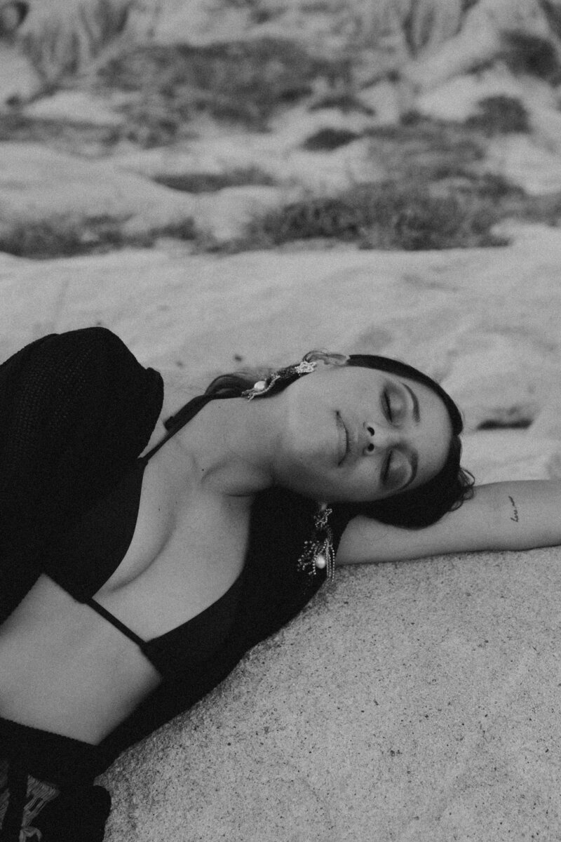 Black and white image of brunette woman in a black bikini top and earrings laying on the beach