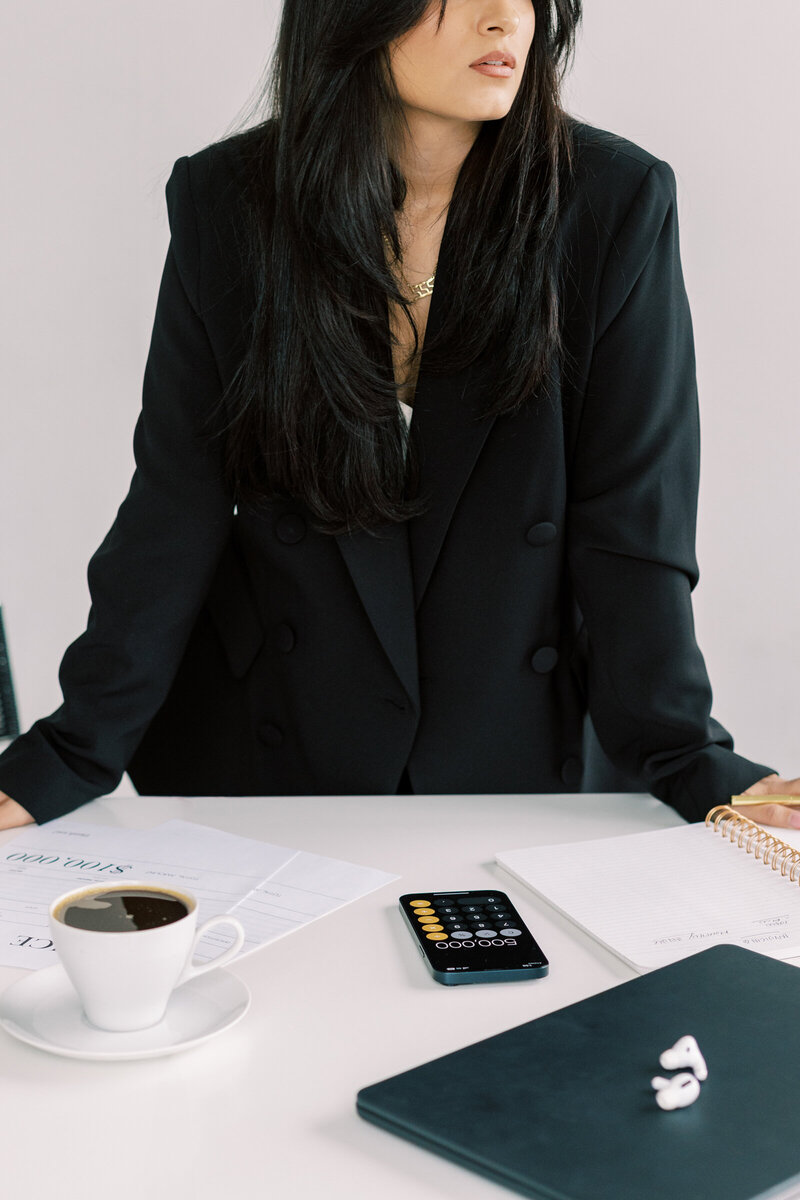 A woman in a black blazer standing at a table with a cup of coffee and calculator 