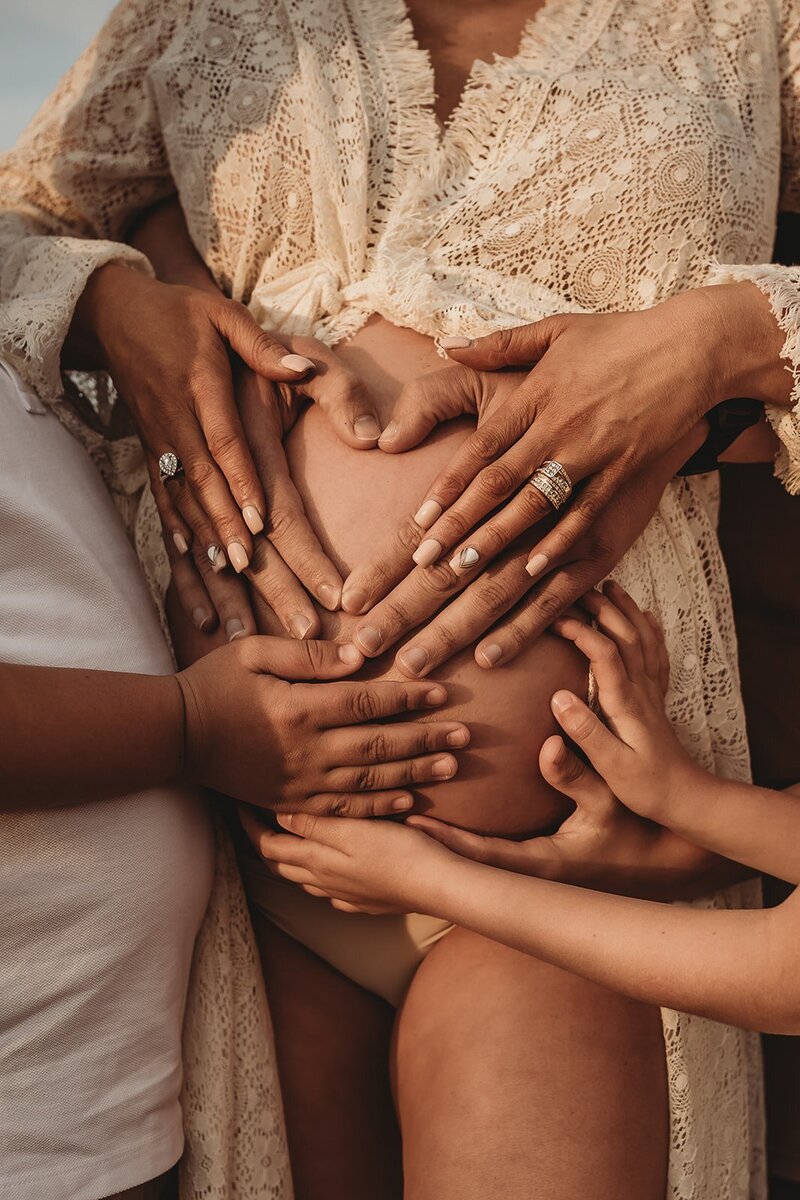 Multiple hands forming heart shapes on a pregnant woman's belly in a lace dress, captured beautifully by an Illawarra maternity photographer.