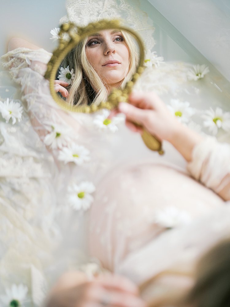 A pregnant mom sits in a bath in a lace dress holding a gold mirror during her maternity photos.