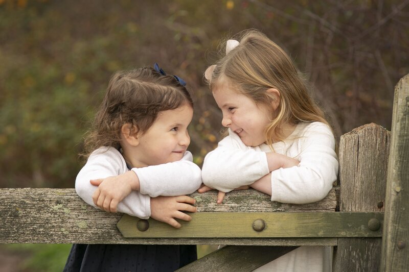 Two young girls in matching cream sweaters lean over a rustic wooden gate, smiling at each other with joyful expressions. The background features soft, natural greenery in an outdoor setting