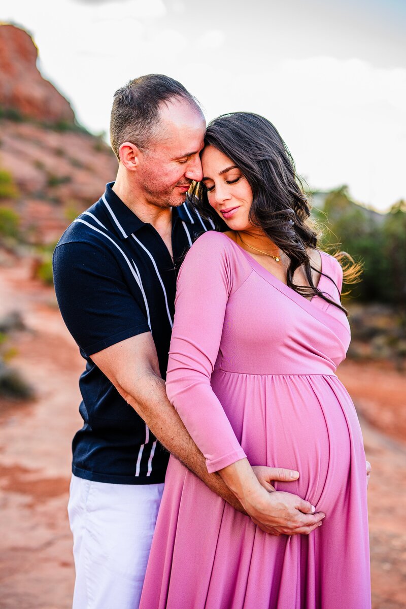 husband holding his wife from behind both hugging her pregnant belly with red rock background in Sedona