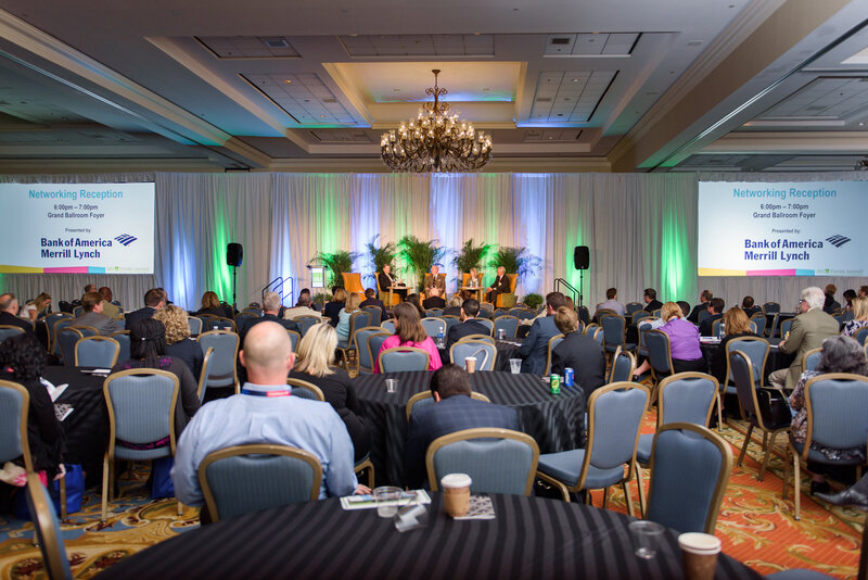 Conference attendees sit at large round tables at a ballroom with a stage and uplighting
