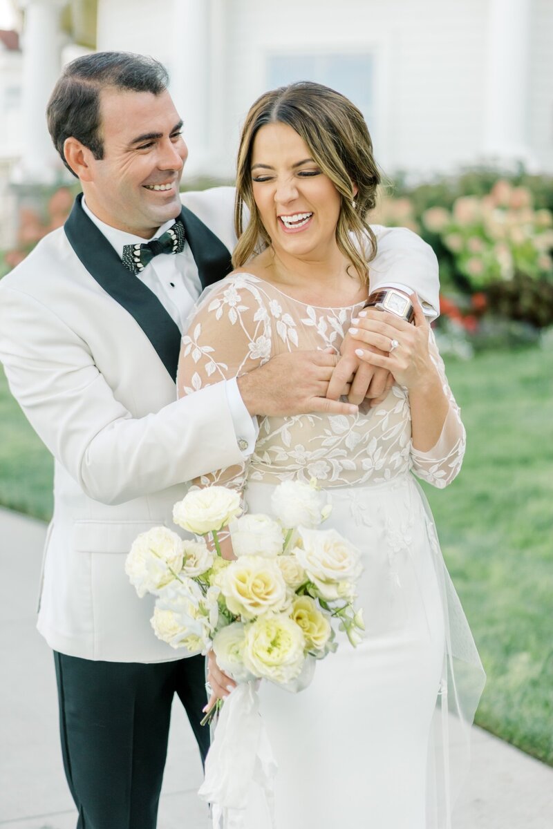 Groom and bride laughing together outdoors, holding a bouquet of white and cream flowers.