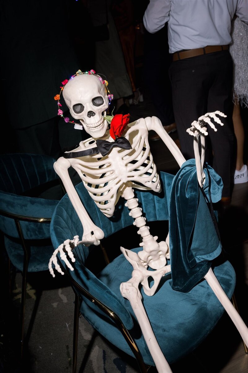 Life size skeleton sitting on a teal velvet chair with a black bowtie on its neck, faux floral led crown on its head, and teal velvet napkin draped on its arm with a red rose in its mouth
