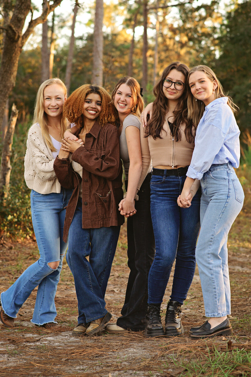 group of girls in jeans standing in a park embracing each other and smiling. 