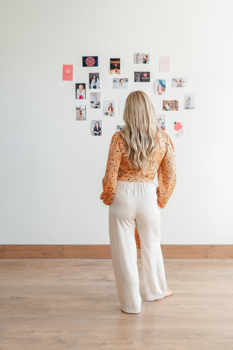 Storycatcher photographer Kristen standing in front of mosaic photo wall back to camera with hands in pockets of cream pants
