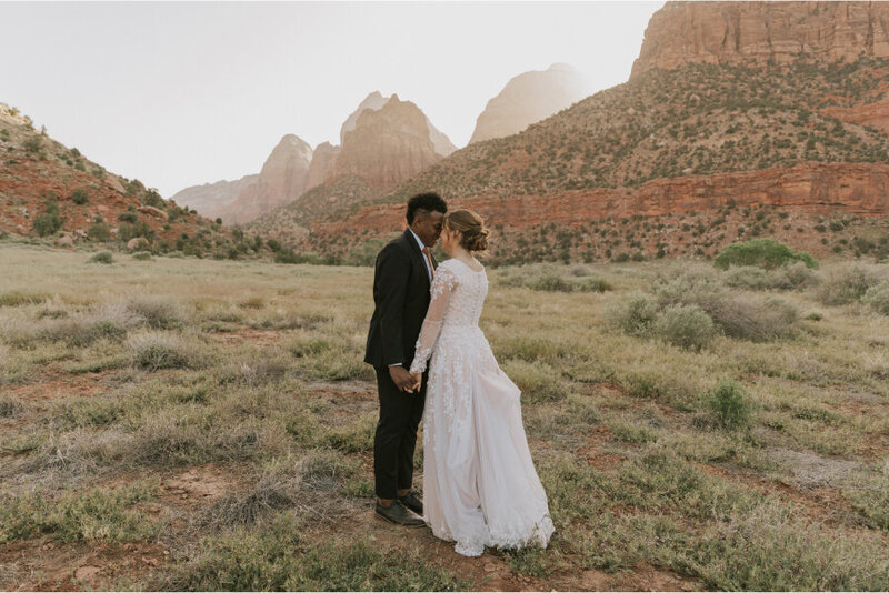 A bride and groom standing close together with their foreheads touching. They are standing in a field of grass and sagebrush with  layered rocks and buttes behind them for hundreds of miles.