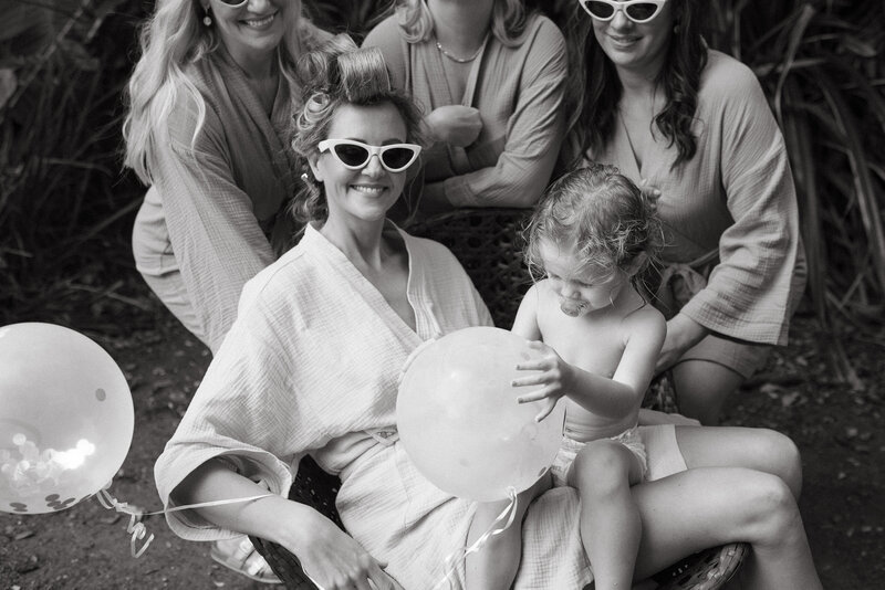 A bride is getting ready with her bridesmaids , they are joyfully holding onto her and her toddler girl , they have vintage glasses and curlers in their hair