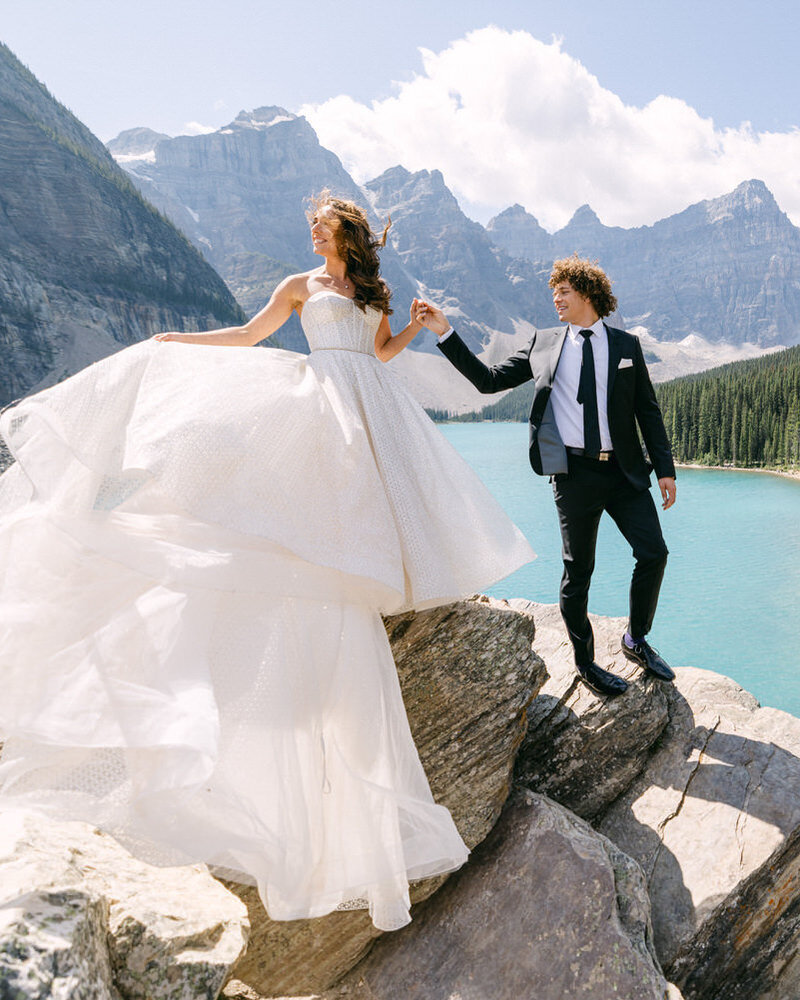 This breathtaking photograph captures a bride and groom holding hands at Moraine Lake, with her dress flowing gracefully in the mountain breeze. Surrounded by towering peaks and the iconic turquoise waters of Moraine Lake, this moment radiates romance and elegance, perfectly framed by the stunning landscape of the Canadian Rockies.