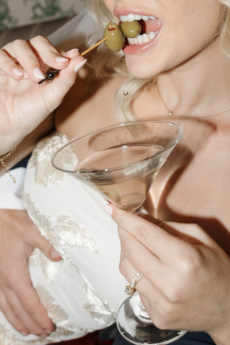 Bride holding a martini glass, biting a green olive showing off her wedding band and ring.