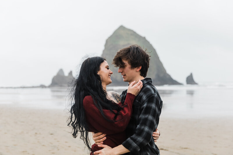A moody engagement session in Cannon Beach Oregon featuring a woman in burgundy smiling at her man wearing a flannel shirt as the rain falls with Haystack Rock in the background. | Erica Swantek Photography