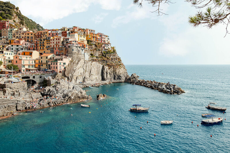 A picturesque coastal village with colorful buildings perched on a rocky cliff overlooking the turquoise sea, reminiscent of serene Caribbean getaways. Several small boats float on the water as people enjoy the beach and shoreline, with sparse trees framing the idyllic scene.