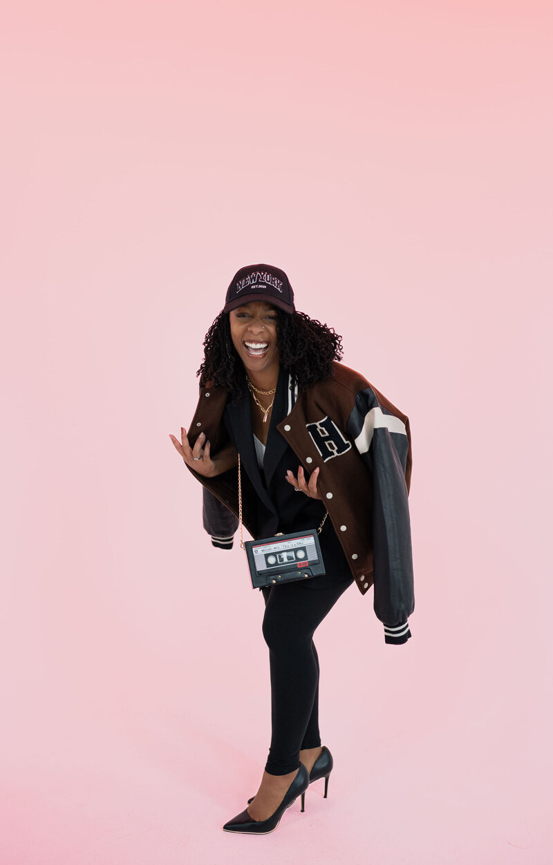 Latesha Lynch, a content strategist, is laughing with a letterman jacket, New York hat, purse shaped like a cassette tape that says 90s Mix, and pumps. 