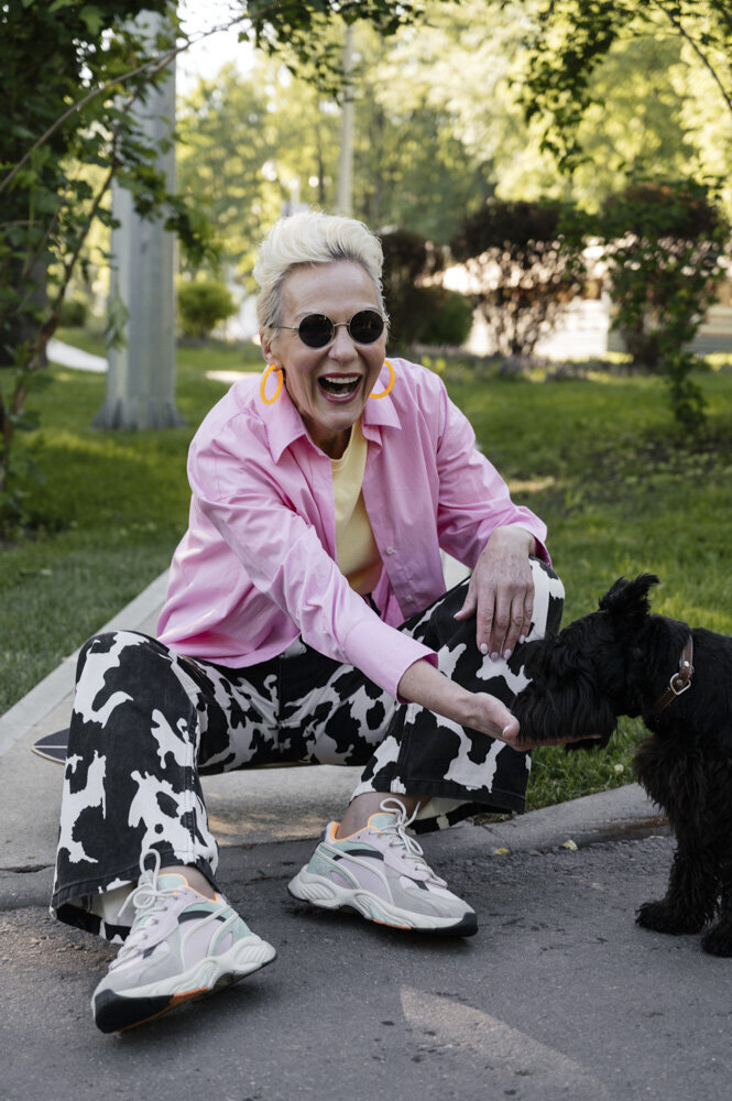 An older white woman wearing a pink button down and cow-print pants sits on a curb outside. She is smiling in the direction of the camera, holding her right hand out to a black shaggy dog.