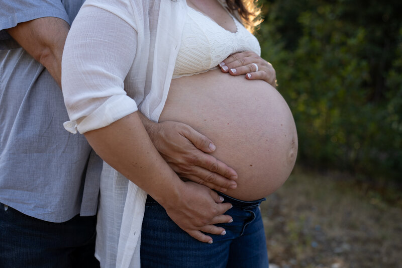 A close up image of a new mother's pregnant belly with her husband's arms hugging her from behind.