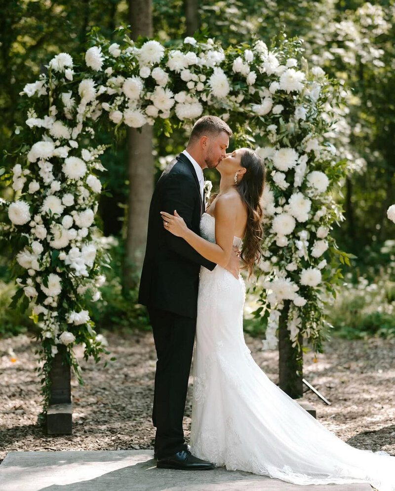 Bride and groom sharing a kiss in front of a lush floral arch during a sunny outdoor wedding ceremony at Bear Ridge Destination.

